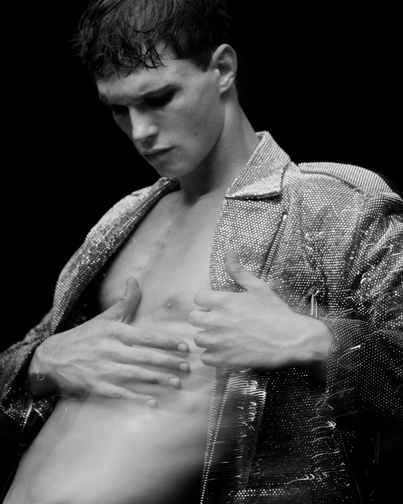 Black and white portrait of a shirtless adult male wearing a sparkling jacket in front of a dark backdrop in a photo studio.
