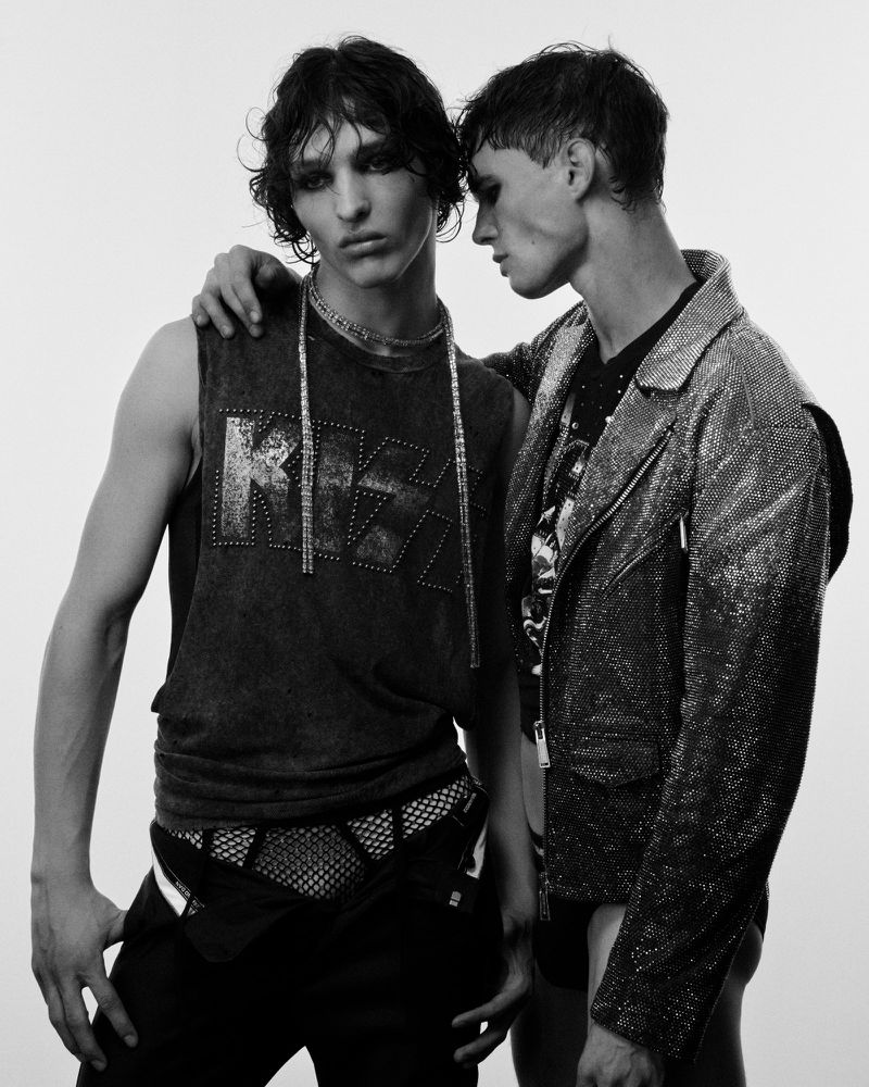 Two young men pose together in a black and white fashion shot, one with a sleeveless shirt with a band logo and the other in a shiny jacket.