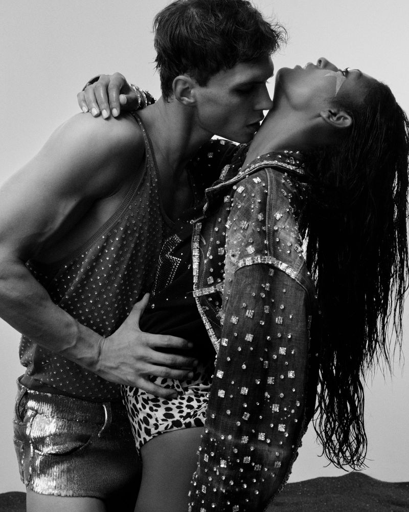 Black and white fashion shot of a shirtless man embracing a woman wearing a studded jacket, with a soft, diffused background.