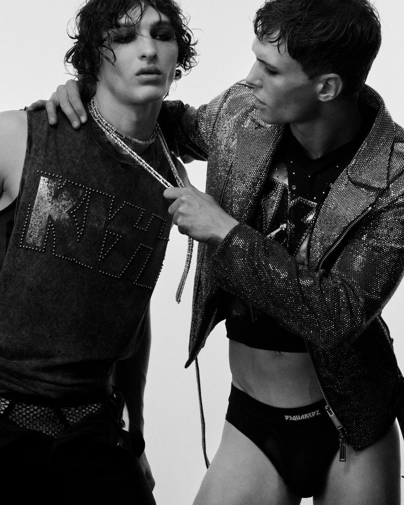 Two men pose indoors, one wearing a KISS graphic tank top with dark makeup, the other in a sequin jacket and briefs, against a white backdrop.