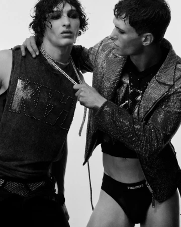 Two men pose indoors, one wearing a KISS graphic tank top with dark makeup, the other in a sequin jacket and briefs, against a white backdrop.