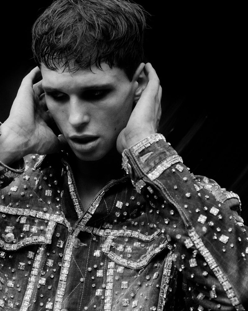 Black and white portrait of a young adult male with dark hair and dramatic eye makeup, wearing an embellished denim jacket.