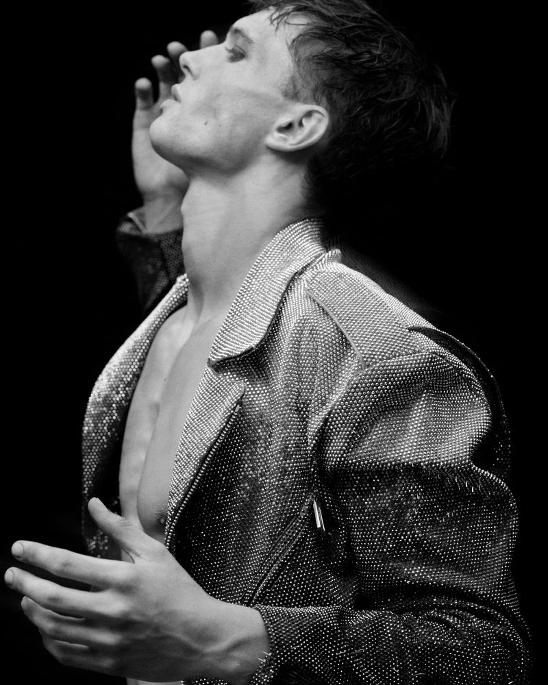 Black and white portrait of a male model wearing a sequined jacket, face tilted upwards with hands raised in a dimly lit photo studio.