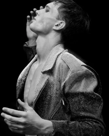Black and white portrait of a male model wearing a sequined jacket, face tilted upwards with hands raised in a dimly lit photo studio.