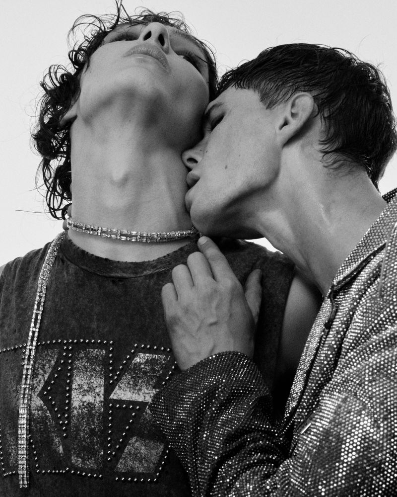 Black-and-white portrait of a male couple in close embrace, one kissing the other's neck, wearing studded fashion tops and jewelry.