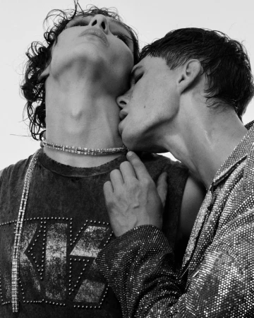 Black-and-white portrait of a male couple in close embrace, one kissing the other's neck, wearing studded fashion tops and jewelry.