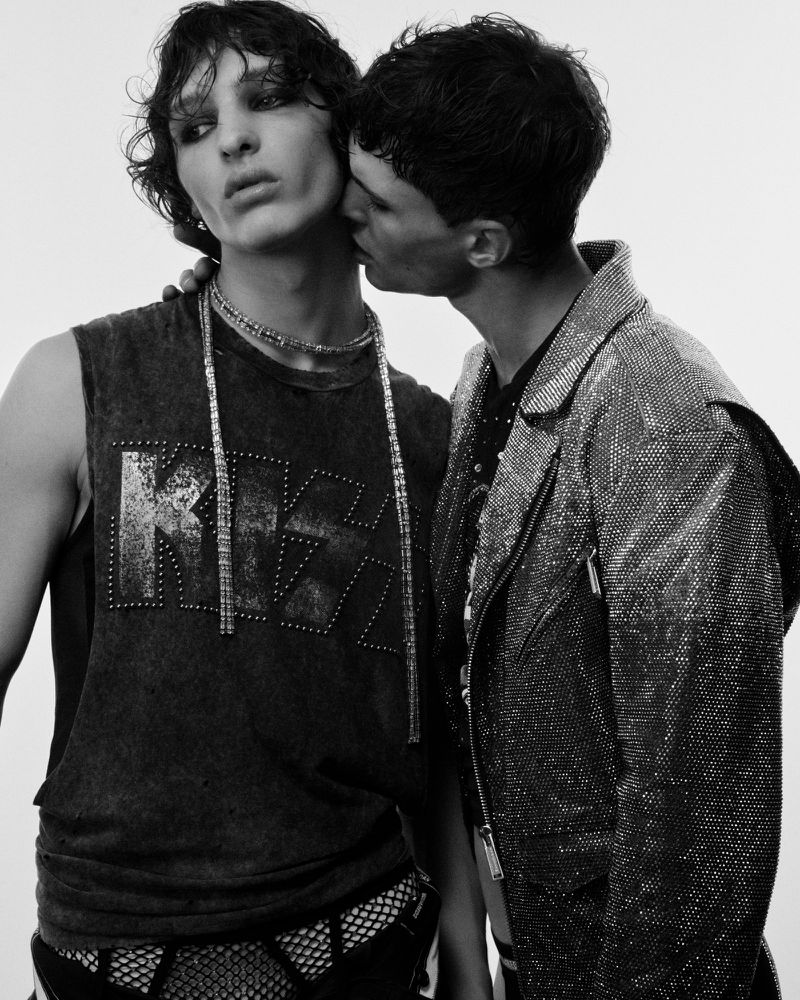 Two young adult men pose closely together in a black and white fashion shot, one with dark hair and smokey eye makeup, the other kissing his cheek.
