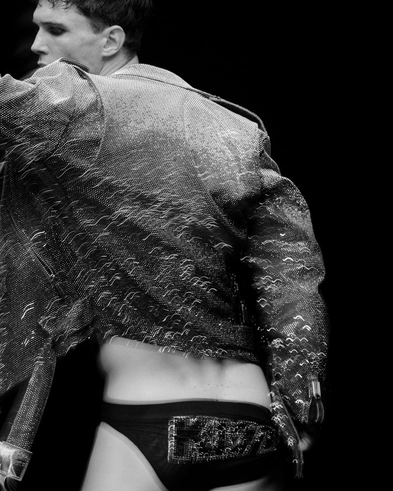 Black and white fashion shot of a slender man in a dark jacket and underwear, posing on a dark background with diffused studio lighting.