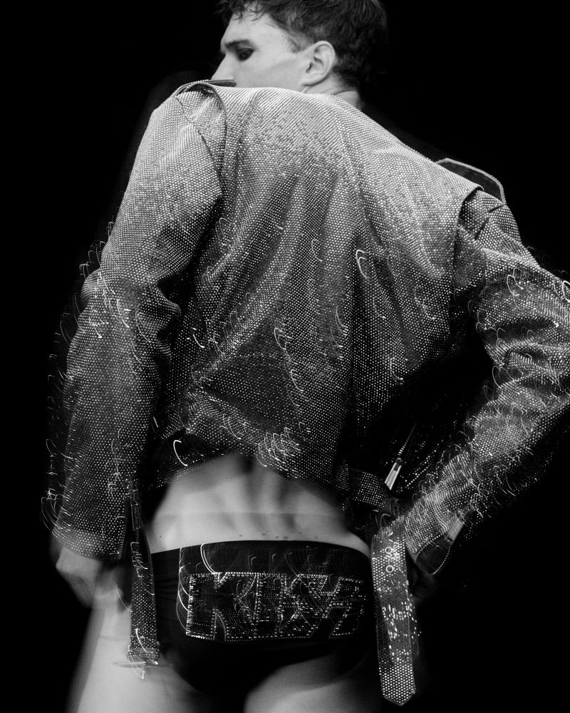 Black and white shot of a person in a jacket and KISS branded underwear standing in front of a black background in a blurred motion.