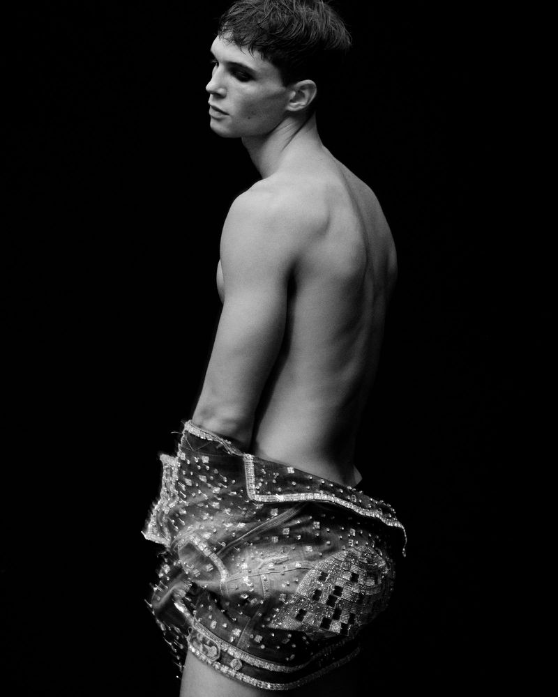 Monochrome portrait of a shirtless adult male model in an embellished jacket, looking away from the camera against a black background.