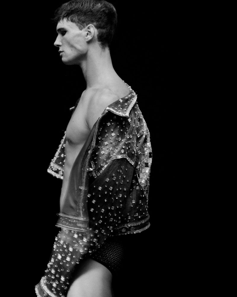 A black and white shot of a shirtless man wearing a sparkly jacket, captured in a studio setting with a black backdrop.