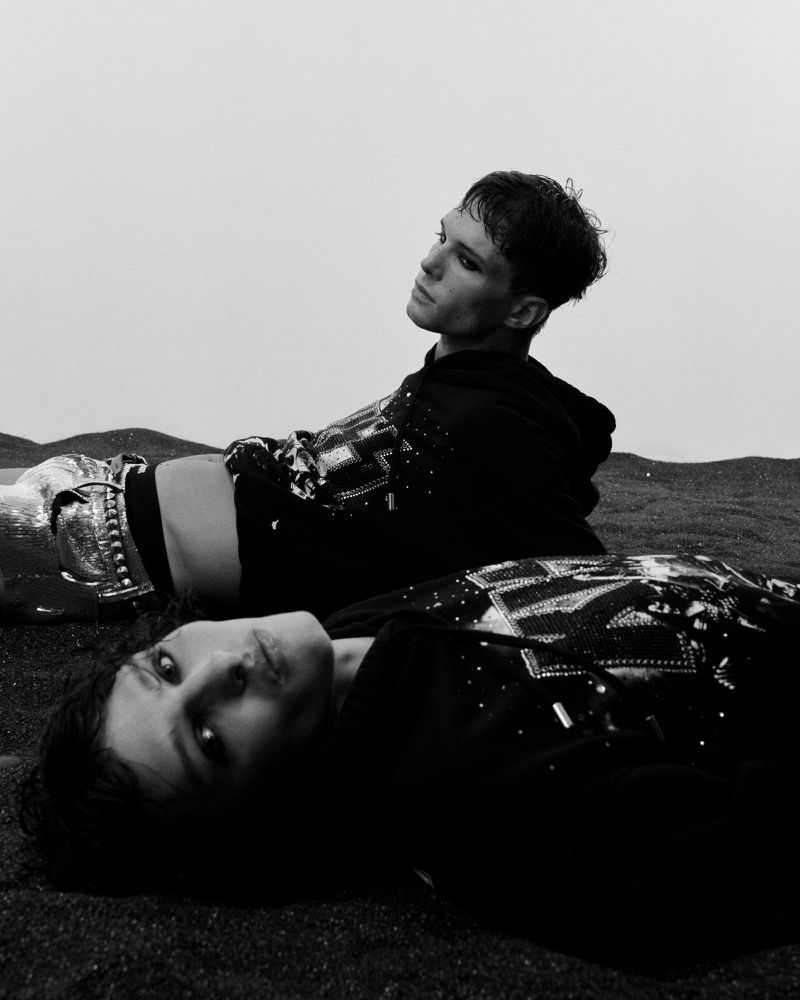 Two young adults lying on dark sand wearing embellished black hooded sweatshirts in a black and white outdoor fashion editorial.