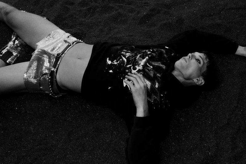 Adult male in sequin shorts and black shirt lies on dark sand. Monochromatic fashion shot with dramatic lighting and shallow depth of field.