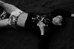 Model Jakob Jokerst lying on dark sand wearing a black graphic sweatshirt and metallic silver shorts.
