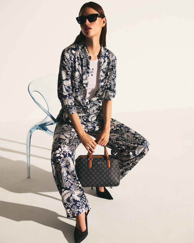 A woman sits on a blue chair in a patterned pantsuit with black shoes, holding a handbag against a white background in a studio setting.
