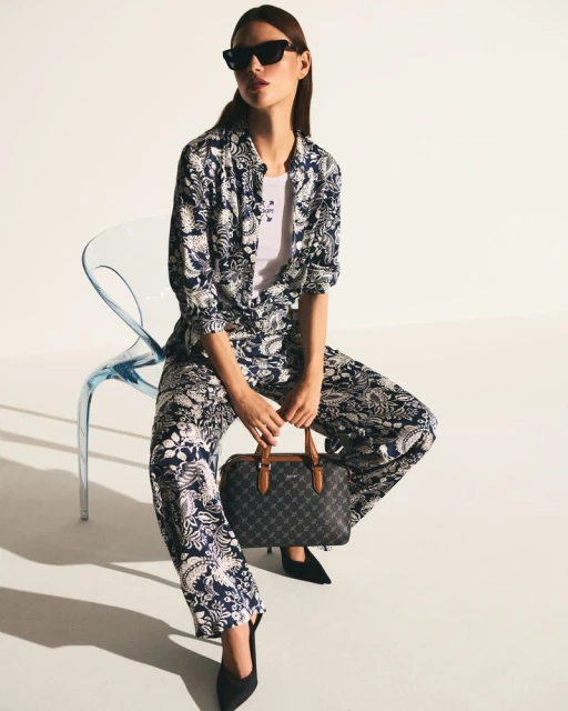 A woman sits on a blue chair in a patterned pantsuit with black shoes, holding a handbag against a white background in a studio setting.