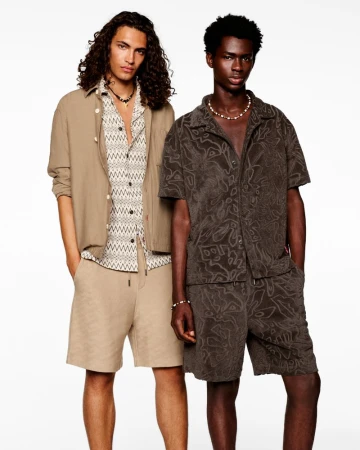 Two male models posing against a white studio background wearing summer patterned shirts and relaxed tailored shorts.