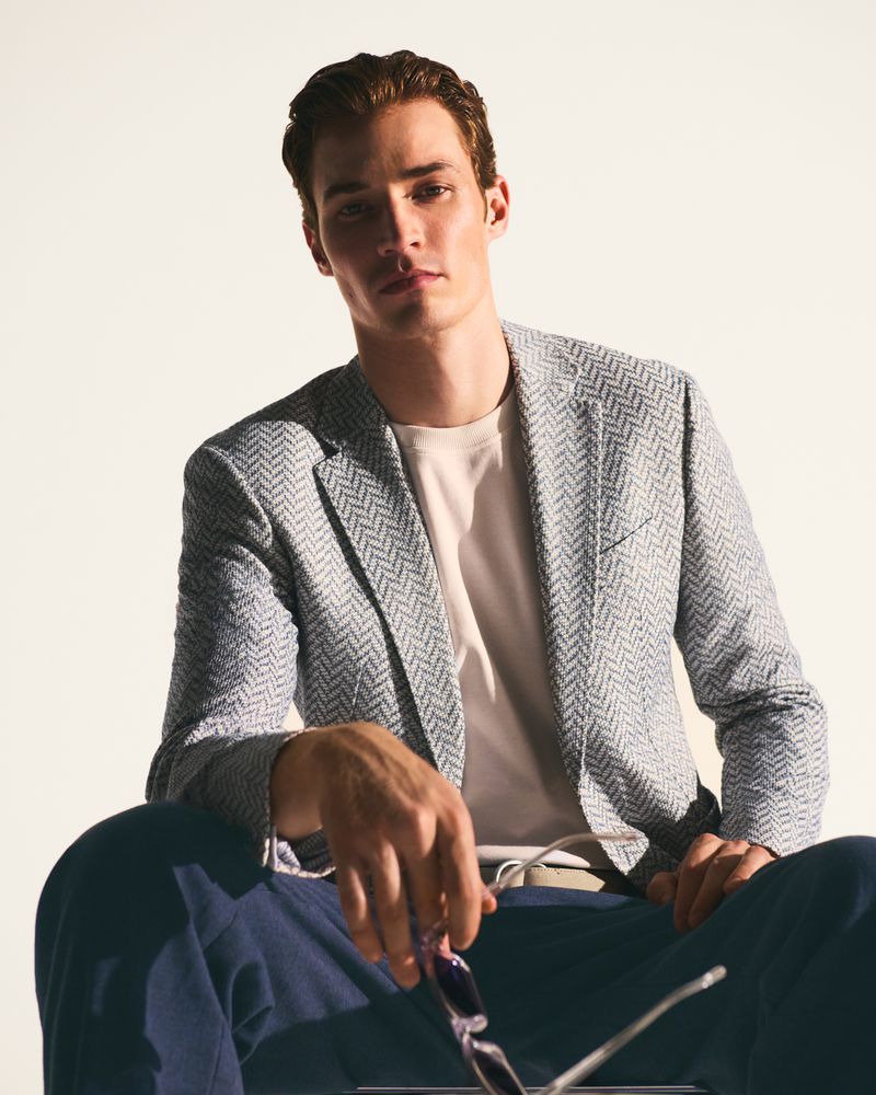 Portrait of a handsome man in a stylish blazer and slacks, sitting against a clean, bright backdrop in a fashion advertisement.