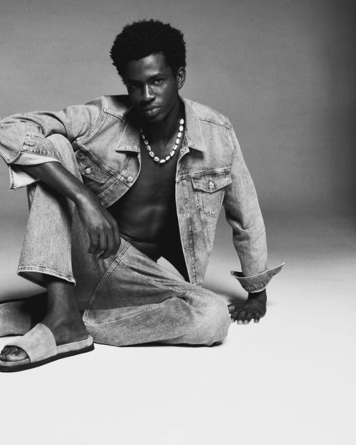 Black and white studio shot of a young adult male wearing a denim jacket and jeans, posing while sitting on the floor. He wears a beaded necklace.