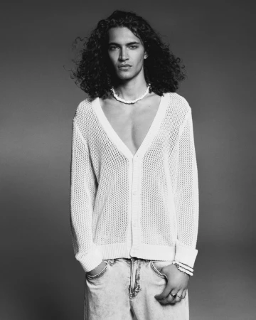 Male model with long curly hair wearing a white open-knit cardigan and light-wash jeans against a smooth grey studio backdrop.