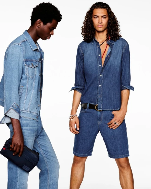 Two adult male fashion models pose indoors wearing denim jacket, denim shorts, and denim shirt against a white backdrop.