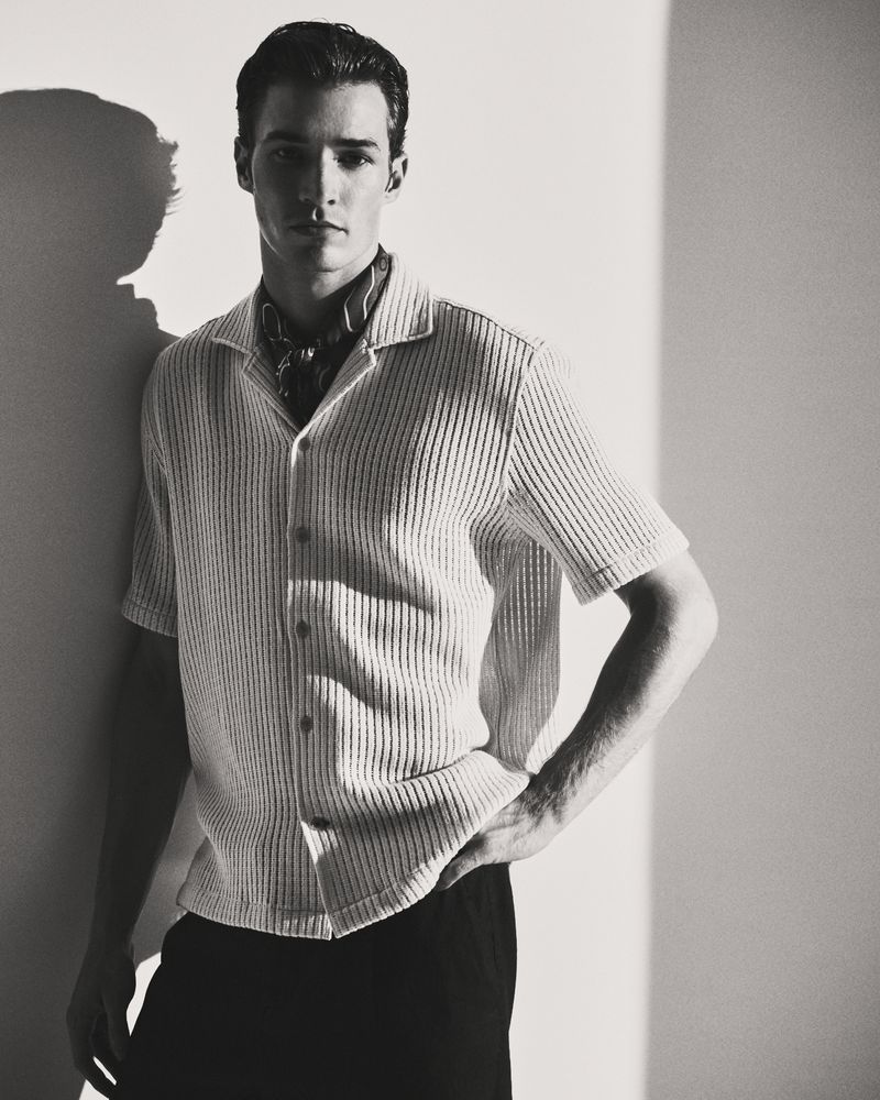 Monochrome portrait of a man in a collared shirt, standing against a wall with shadow, showcasing a blend of fashion and portraiture.