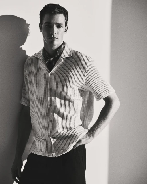 Monochrome portrait of a man in a collared shirt, standing against a wall with shadow, showcasing a blend of fashion and portraiture.