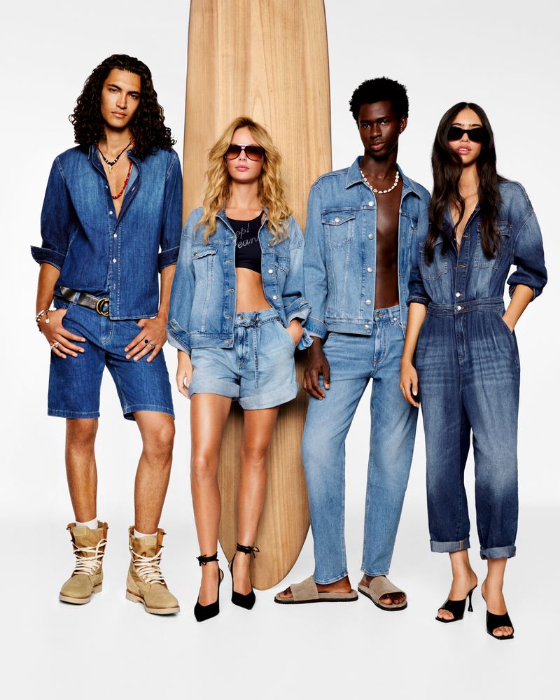 Four models posing in all-denim outfits beside a wooden surfboard against a bright white seamless background.
