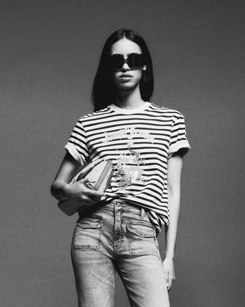 A fashionable young woman in a striped t-shirt and jeans, wearing sunglasses, poses in front of a studio backdrop, holding a clutch.
