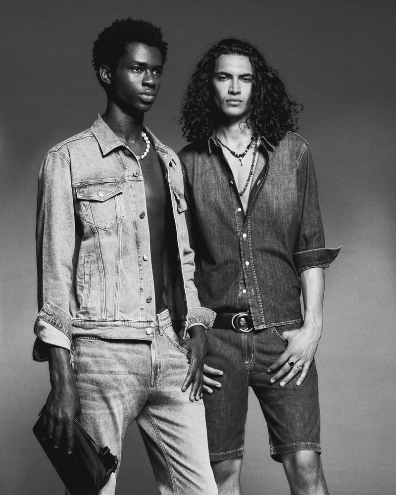 Two male fashion models pose in a studio wearing denim jackets and jeans. One model wears an open denim shirt and dark denim shorts.