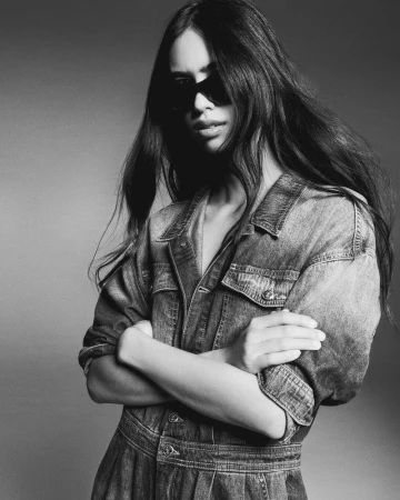 Black and white portrait of a woman wearing dark sunglasses and a denim jumpsuit with arms crossed against a studio backdrop.