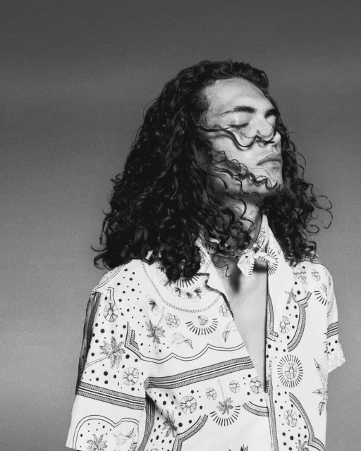 Monochrome portrait of a young adult male with curly hair, eyes closed, wearing an open patterned shirt against a plain background.