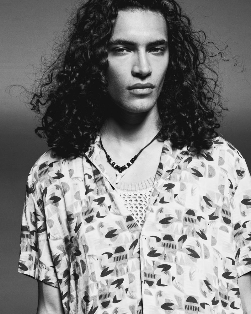 Black and white portrait of a man with long curly hair wearing a patterned short-sleeve shirt against a neutral background. 