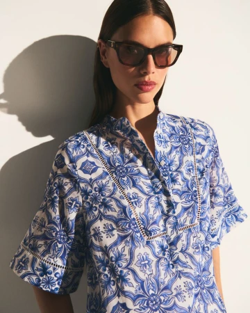 A woman wearing dark sunglasses and a blue and white patterned blouse poses against a white wall with a defined shadow.