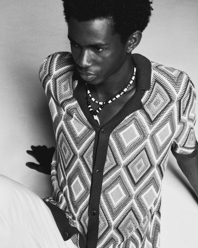 Black and white portrait of a Black male model in a geometric diamond-knit polo shirt and beaded necklace against a plain backdrop.