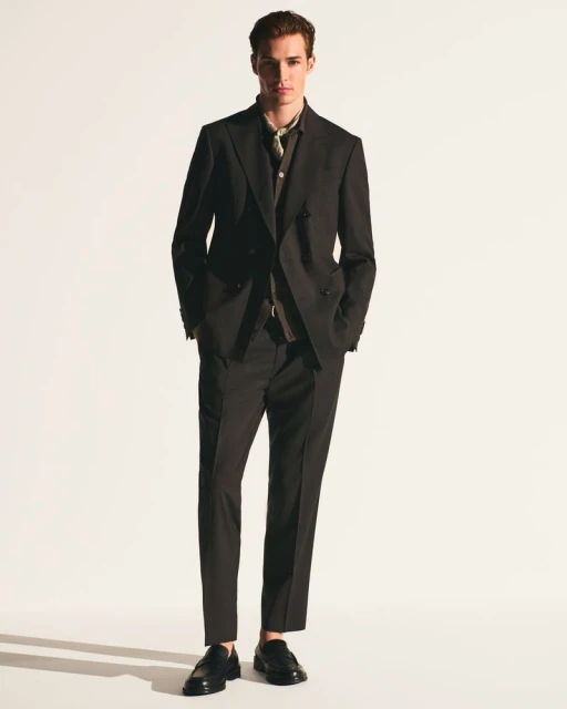 Adult male model poses in a studio wearing a dark suit, a patterned shirt, and black loafers against a plain white backdrop.