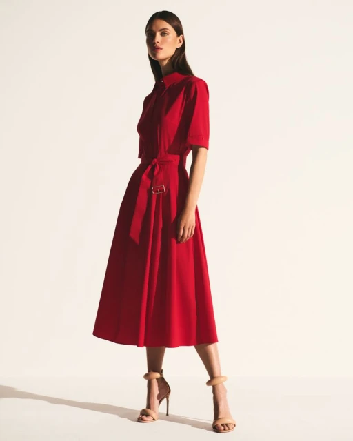 Woman in red midi dress with a-line skirt, belt, and short sleeves, standing in a photo studio with neutral-color stiletto heels.