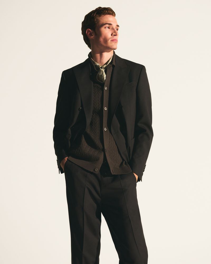 Male model wearing a tailored dark suit jacket, buttoned knitwear, and a patterned neck scarf against a bright studio background.