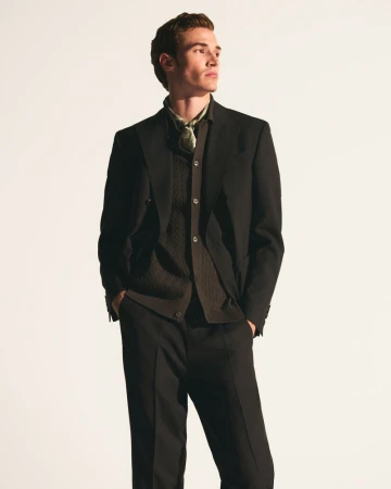 Male model wearing a tailored dark suit jacket, buttoned knitwear, and a patterned neck scarf against a bright studio background.