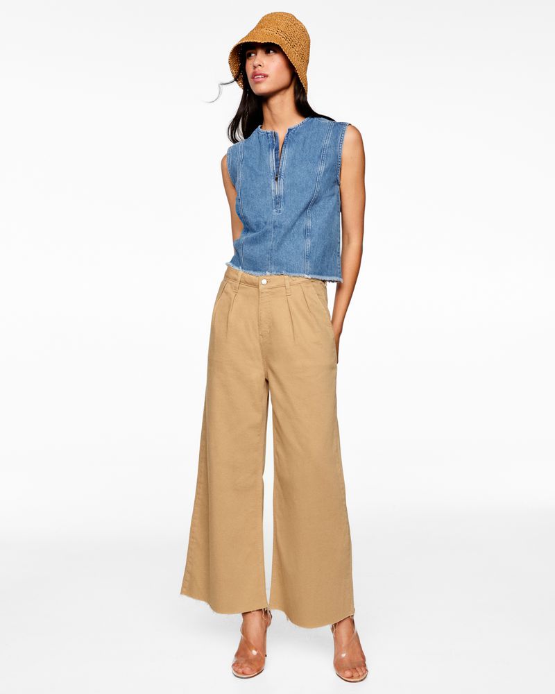 Woman in a denim top, wide-leg tan pants, woven bucket hat, and high heels stands against a white backdrop in a full-length studio shot.