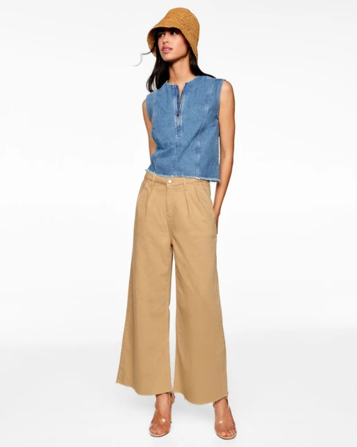 Woman in a denim top, wide-leg tan pants, woven bucket hat, and high heels stands against a white backdrop in a full-length studio shot.