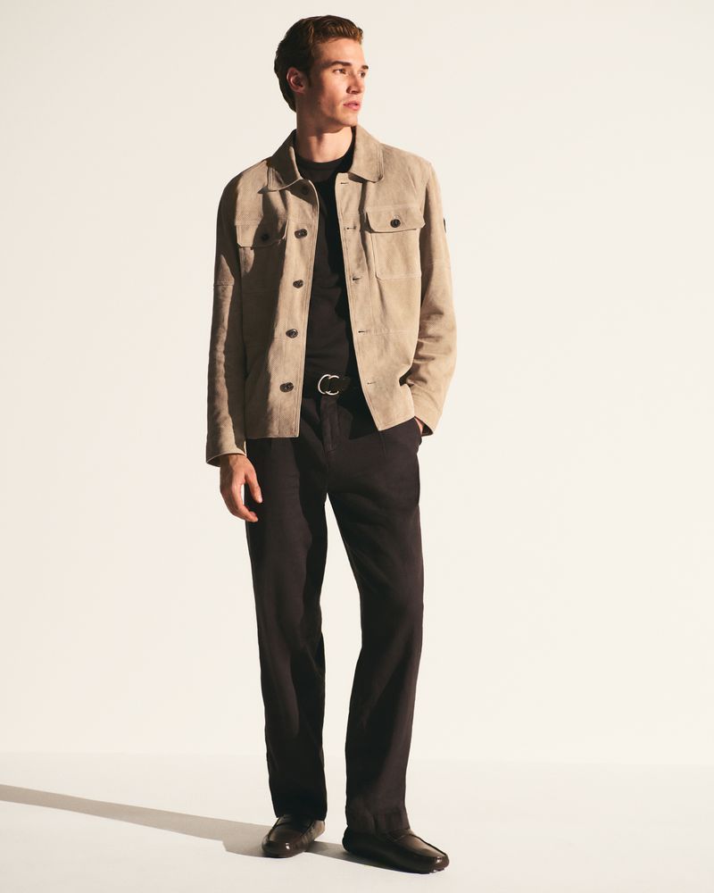 A fair-skinned adult man in a beige jacket and black trousers poses against a stark white backdrop in a fashion-style, full-body shot.
