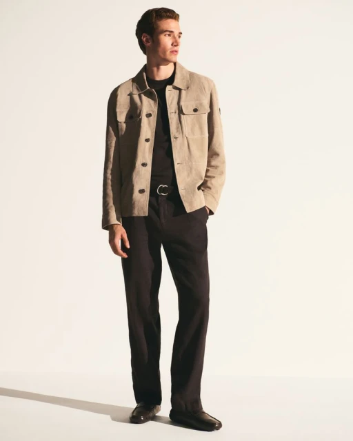 A fair-skinned adult man in a beige jacket and black trousers poses against a stark white backdrop in a fashion-style, full-body shot.