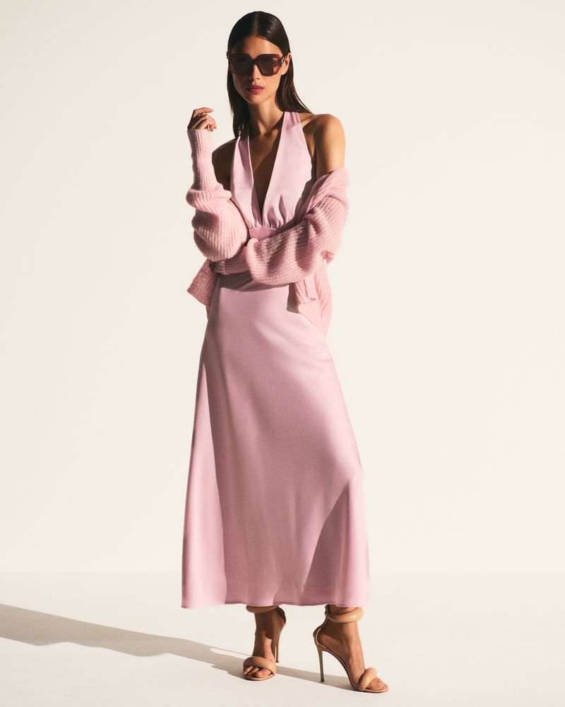 Model in a pastel pink halter dress, matching cardigan, oversized sunglasses, and nude stiletto sandals against a white background.