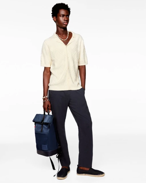 A young Black man stands in a photo studio wearing a cream knit shirt, navy pants, and espadrilles while holding a navy backpack.