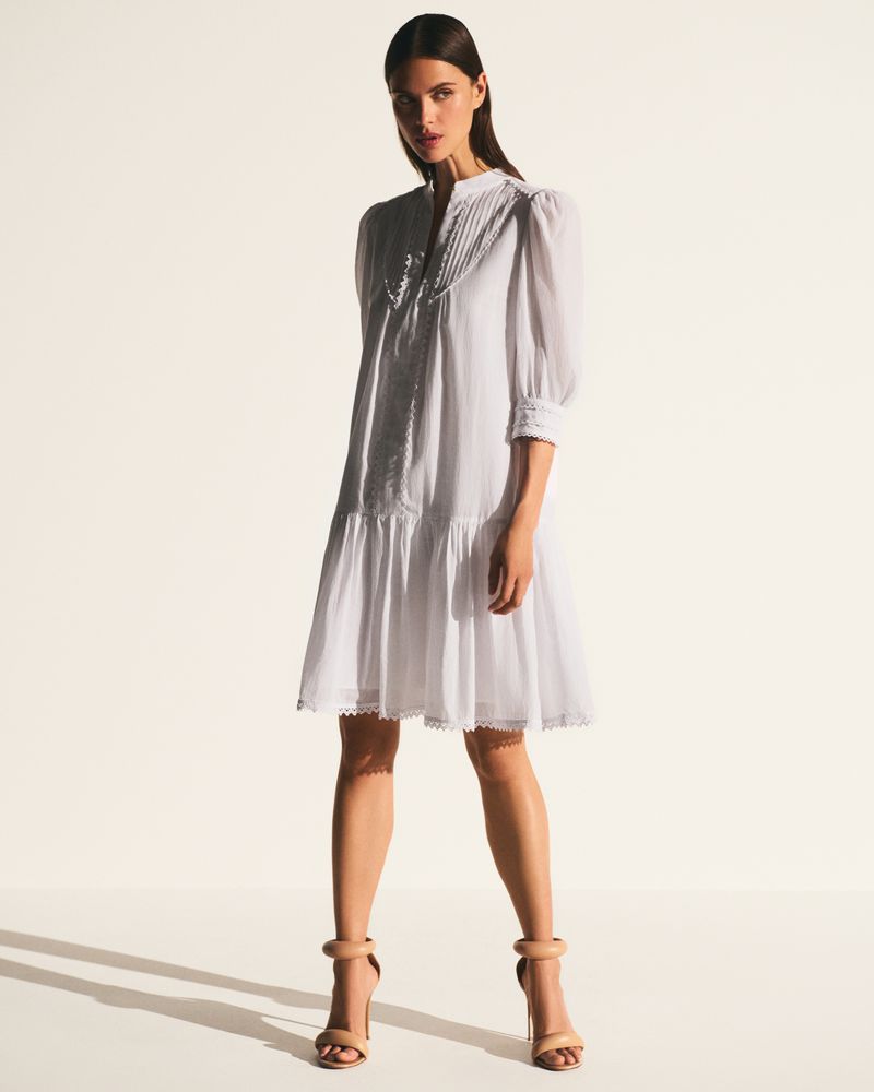 A full shot of a woman posing in a white dress with puff sleeves and nude heels against a bright, neutral backdrop.