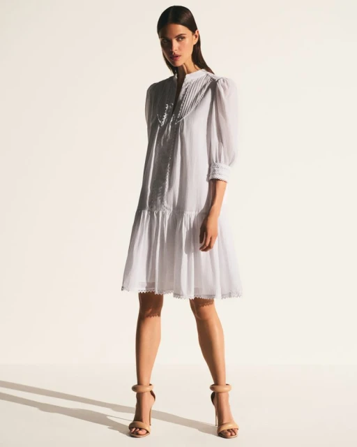 A full shot of a woman posing in a white dress with puff sleeves and nude heels against a bright, neutral backdrop.