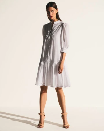 A full shot of a woman posing in a white dress with puff sleeves and nude heels against a bright, neutral backdrop.
