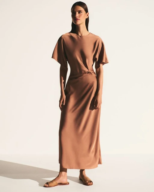 A tanned-skin adult female model poses in a full shot wearing a tan midi-length dress with short sleeves, and flat sandals.