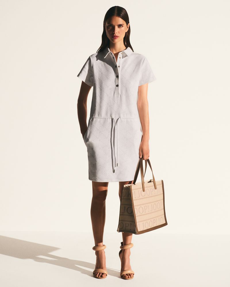 Brunette fashion model wearing a white collared mini-dress, high heel sandals and a tan tote bag posing in a bright photo studio.